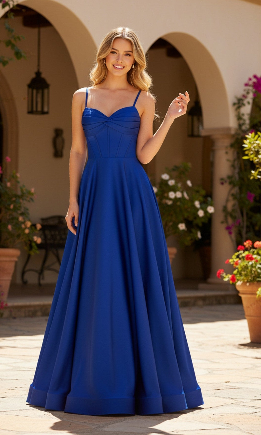 A woman wearing the PromGirl Exclusive 250235 long prom dress in royal stands outdoors in a courtyard with potted plants and arched architecture in the background.